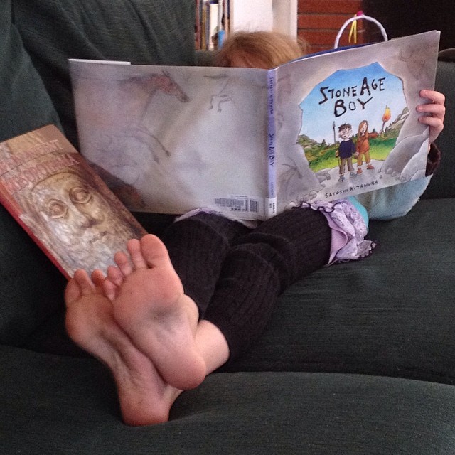 a small barefoot child stretched out on a sofa, reading a picture book