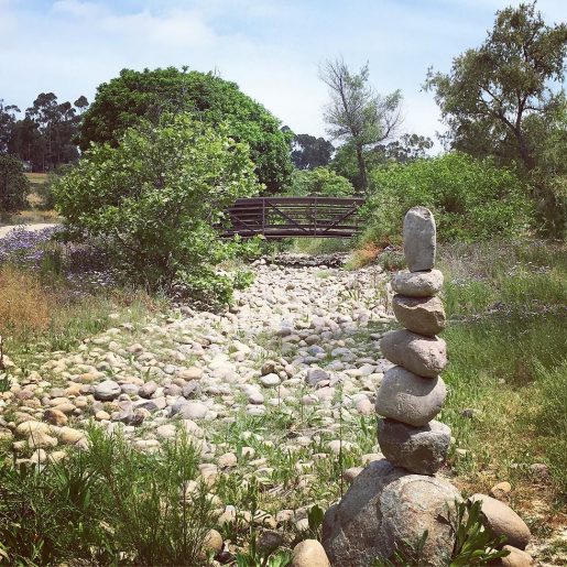 A tower of small stones at the side of a dry, rocky creekbed, with a small brown footbridge in the distance.